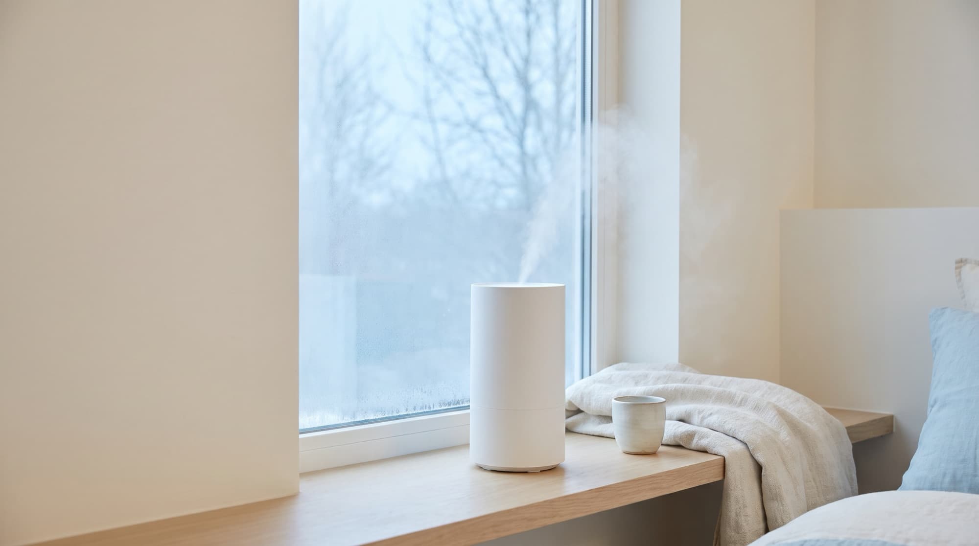冬の朝の寝室。窓辺の木の棚に白い加湿器と陶器のカップが置かれ、加湿器から白いミストが立ちのぼっている